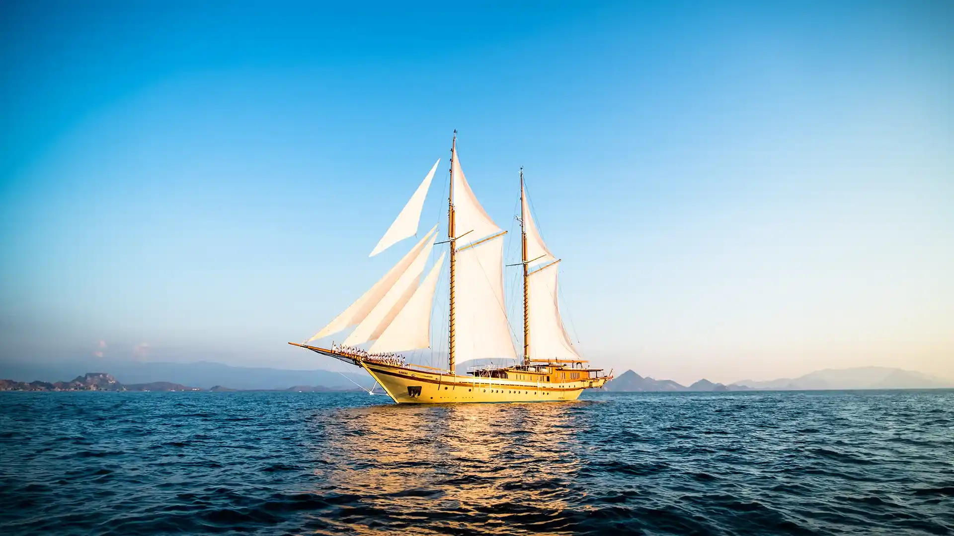 Phinisi liveaboard under full sail among Komodo's islands at golden hour